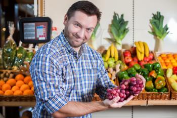 Portrait of happy man in shirt showing a bunch of grapes in supermarket. Boxes with fruits and vegetables on the background. 