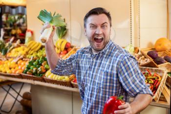 Man jokes with leek and red pepper in hands.  Boxes with fruits and vegetables on the background. Marketplace.