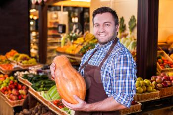 Smiling man in apron offers fresh vegetables in grocery shop.