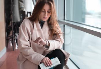 Young woman in international airport, waiting for her flight, checking her wrist watch and looking upset or worried. Arrival, missed, canceled or delayed flight concept.