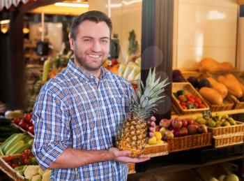 Portrait of smiling man in shirt holds ripe pineapple in hands. Grocery on the background.