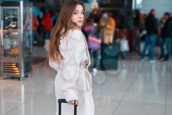 Young woman in white coat at the modern airport, people traveling with luggage. Queue for registration on the background.