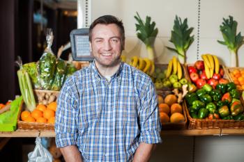 Portrait of smiling man in shirt selling vegetables in a shop. Boxes with fruits and vegetables on the background