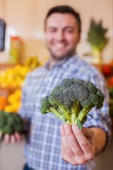 Smiling man holding broccoli at arm's length. Supermarket on the background.