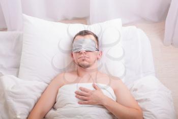 Sleeping young man in sleep mask on bed with big pillow. Comfort sleeping concept. Insomnia problem solution.