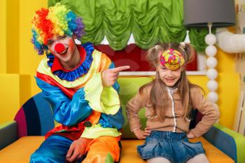Little girl has put on a cap, and the clown laughs at her. Kindergarten on the background.