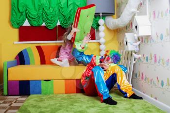 Happy little girl beats clown with a big pillow. Playroom with colourful sofa on the background.