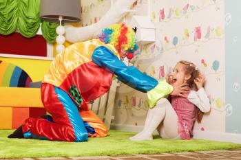 Frightened girl and a clown in colorful costume playing on the green carpet.