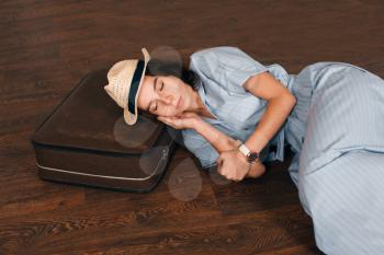 Young woman in light blue dress and white hat put her head on suitcase and sleeps on the floor. Waiting concept.