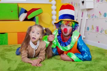 Smiling child and the clown lie on a carpet in the nursery. Colourful sofa, nesting box and fence on the background. 
