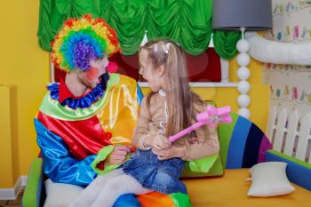 Little girl sitting on the lap of a cheerful clown with funny makeup. Children's friendship forever.