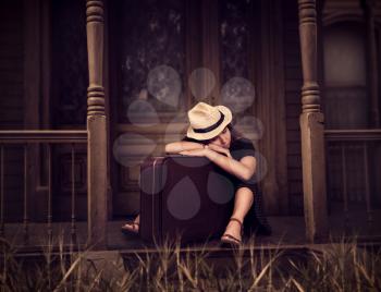 Portrate of young woman in white hat with retro suitcase is sitting on threshold of old house.