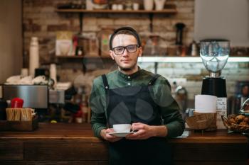 Waiter in eyeglasses with a cup of coffee in hands has leaned the elbows on a bar counter.