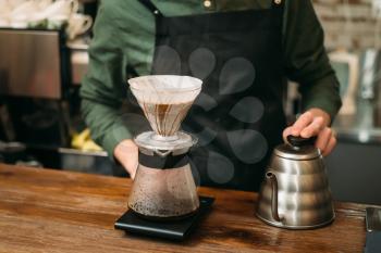Metal coffee pot and glass on a bar counter against waiter in black apron. Blury coffee house on background.