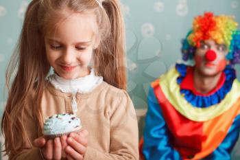 Smiling little girl holds cake in hand and makes a wish. Funny clown on the background.