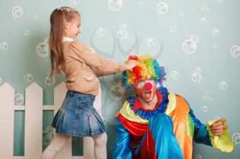 Little girl pulls clown hair. White fence decoration on the background.