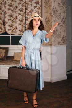 Young girl standing in the room with suitcase and holds out a hand. Vintage travel waiting concept. Sofa and window with curtains on the background.