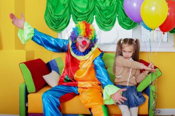 Birthday celebration with clown in amusing costume. Little girl holds colorful air balloons in her hand.
