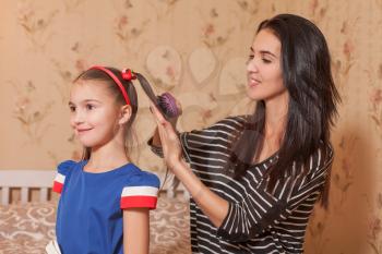 Young mother making hairstyle with hairbrush to her little daughter 
