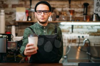 Barman in black apron stretches plastic cup of coffee in hands.