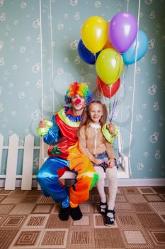 Happy little girl with the clown shake on a swing. Clown hold air balloons in his hands.