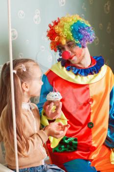 Joyful little girl with happy clown sitting on a swing, he invites her to eat birthday cake
