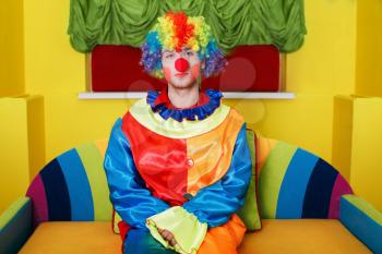 Clown in rainbow colored costume with makeup and red nose sitting on colorful sofa.