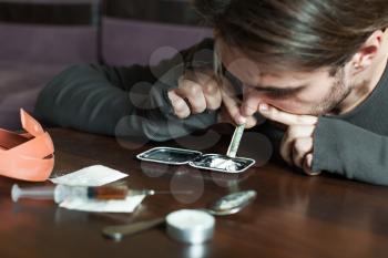 Addict man smells cocaine from a mirror. Drugs on wooden table.