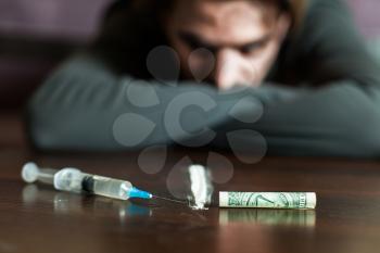 Addiction man look at the table with syringe, dollars and cocain trace. Abuse of narcotics leads to a depression.