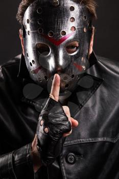 Serial maniac in hockey mask and cutted leather gloves show do not talk sign leaning his index finger to his lips. Black background