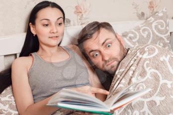 Young woman with book and man under blanket are lying in bed. Couple reading a book before sleep.