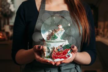 Glass christmas snowball in female hands closeup. Woman in grey apron with decorative glass globe in hands.