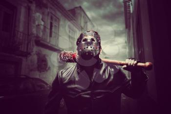 Angry psycho man in hockey mask and black leather coat with bloody baseball bat with a chain wrapped around. Maniac waiting for his victim on night city street