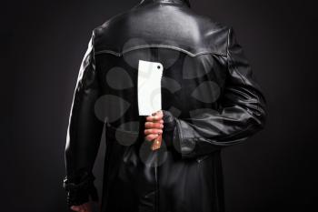 Serial murderer in black leather coat holding meat cleaver behind the back, black background
