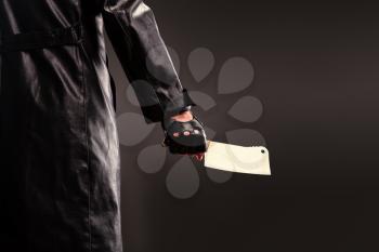 Killer in black leather coat holding meat cleaver in hand against black background.