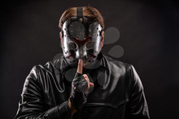 Serial maniac in hockey mask and cutted leather gloves show do not talk sign leaning his index finger to his lips. Black background