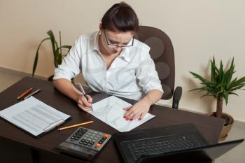 Female bookkeeper in glasses working at the table with laptop, calculator and accounting documents