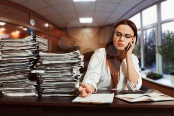 Young woman bookkeeper using mobile phone at workplace, stacks of documents on the table