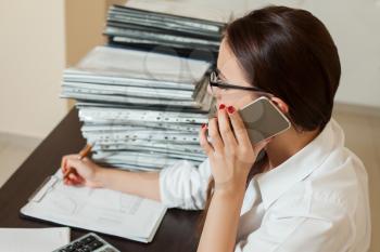 Young female bookkeeper in glasses talking by phone, stacks of documents on the table