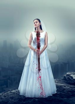 Young bride in white wedding dress with bloody baseball bat, foggy city on background