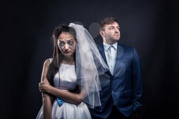 Disheveled bride with tear-stained face and brutal groom in suit, studio photo shoot, black background.