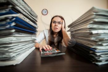 Doomed female accountant against big stacks of documents and calculator