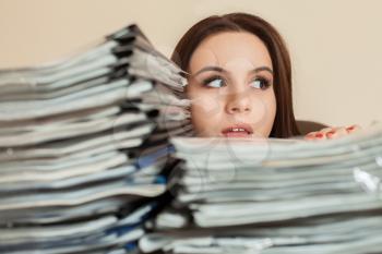 Young female bookkeeper looking out of the large stacks of documents