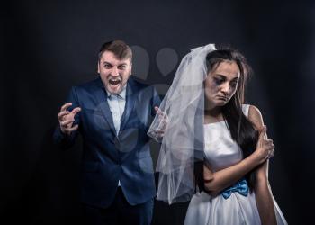 Bride with tearful face and terrible brutal groom in suit, studio photo shoot, black background.