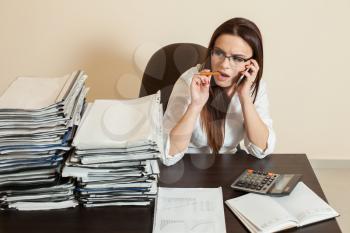 Young female accountant in glasses using cell phone at workplace, stacks of documents on the table