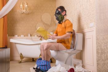Man in gas mask with pants down sitting on the toilet and reading a book