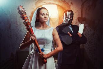 Newlyweds maniacs, an interior of old prison on background. Couple with bloody bat and meat cleaver, groom in hockey mask