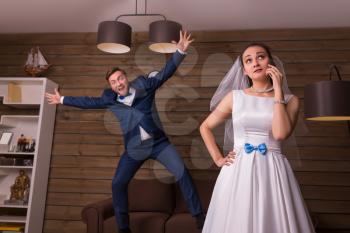 Portrait of young bride with mobile phone against playful groom, wooden room on background.