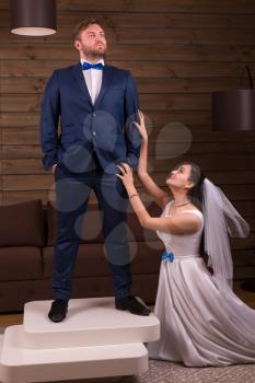 Beautiful bride in white dress and veil makes marriage offer to the groom in suit standing on table, wooden room interior on background