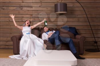 Drunk bride with bottle of alcohol and groom sleeping on couch after wedding celebration. Wooden interior of the room on background